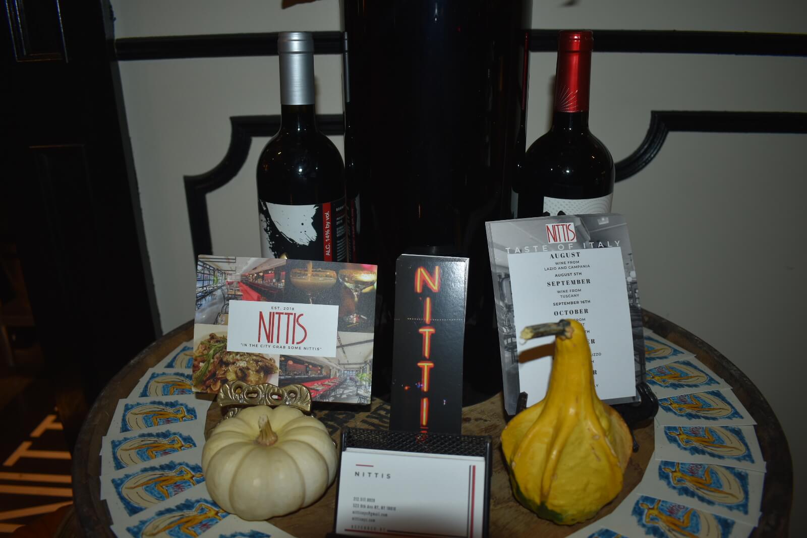 Nittis business cards displayed on a table in front of two bottles of wine and behind a miniature pumpkin and gourd