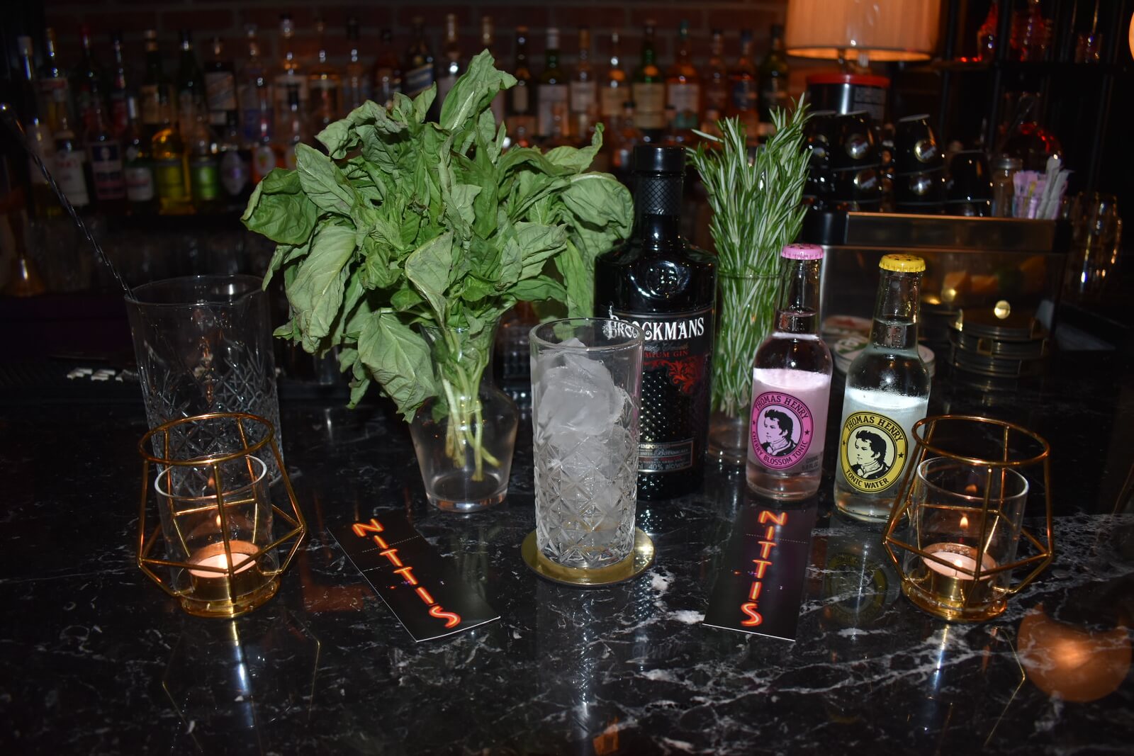 A glass of ice in front of a bottle of Gin and Tonic Water with glasses of garnish